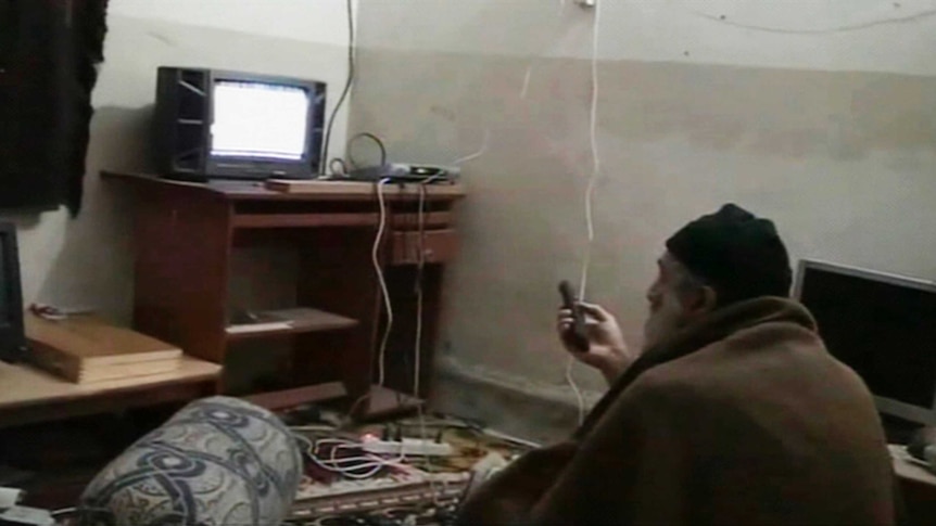 Osama bin Laden wearing a beanie and wrapped in a blanket sits on the floor watching TV.