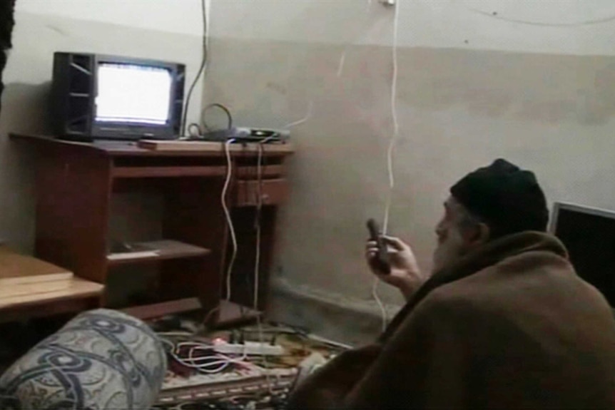 Osama bin Laden wearing a beanie and wrapped in a blanket sits on the floor watching TV.