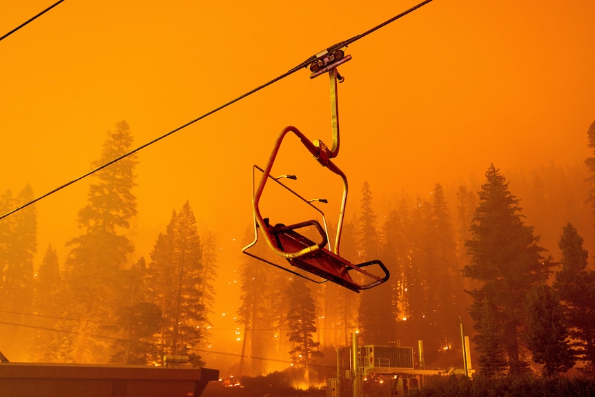 A chairlift dangles overhead as pine trees are engulfed by fire behind it. 