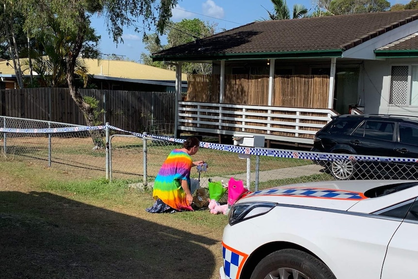 A woman kneels down outside the one-story home at its wire fence, laying a bunch of flowers.