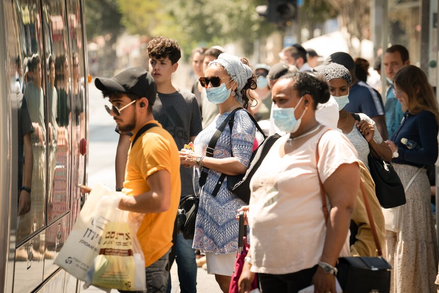 A group of people wearing masks and holding bags line up at a bus stop. 