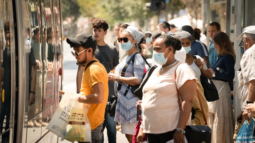 A group of people wearing masks and holding bags line up at a bus stop. 