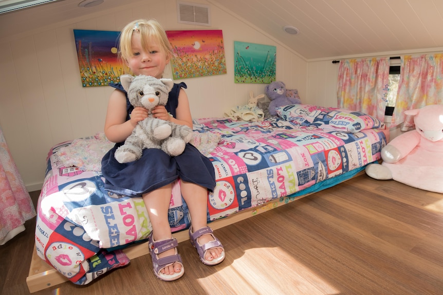 A little blond girl is sitting on her bed in a small space with colorful pink toys and drawings around her.