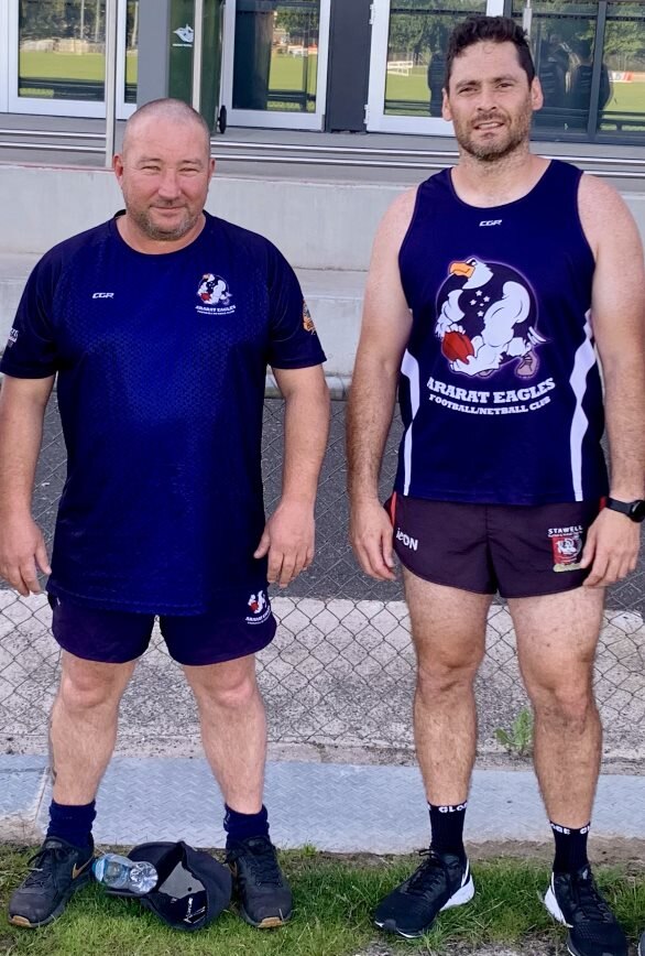 An older bald man in blue and black t-shirt with logo, stands next to a younger man in an Ararat Eagles jersey. Both serious.