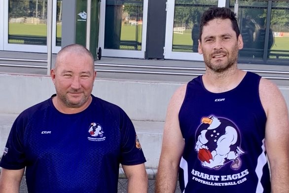 An older bald man in blue and black t-shirt with logo, stands next to a younger man in an Ararat Eagles jersey. Both serious.