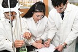 Three students in white lab coats write notes and observe science experiment paraphernalia on a desk.