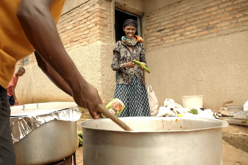 A woman holds a vegetable in her hand smiling as someone is cooking in front of her