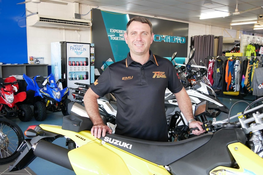 Middle-aged man with short dark hair standing behind a motorcycle in a black polo shirt.