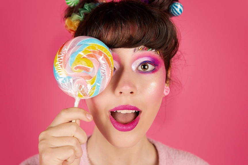 A woman wearing pink makeup and holding a lollypop over one eye