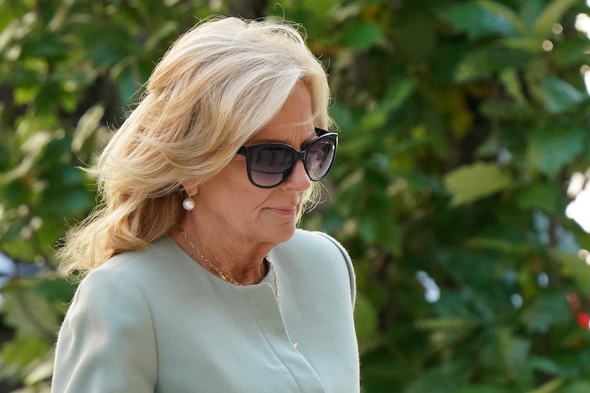 Jill Biden, wearing sunglasses and a pale green top, walks in front of a leafy backdrop.