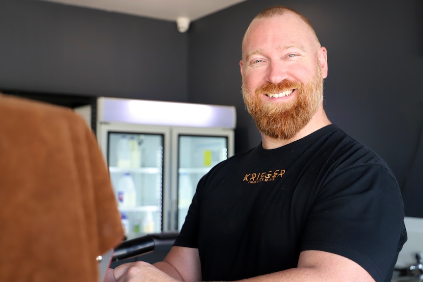 man with beard and balding hair smiles in front of coffee machine