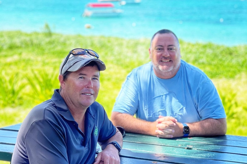 Anthony Riddle and Christian Young sit on a park bench in front of crystal clear, blue water.  
