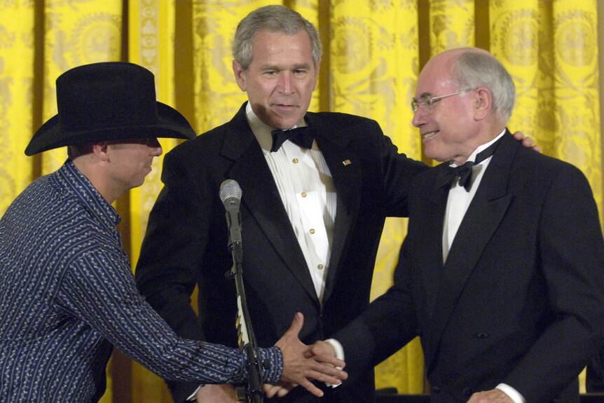 Two men in tuxedos shake hands with a man in a cowboy hat