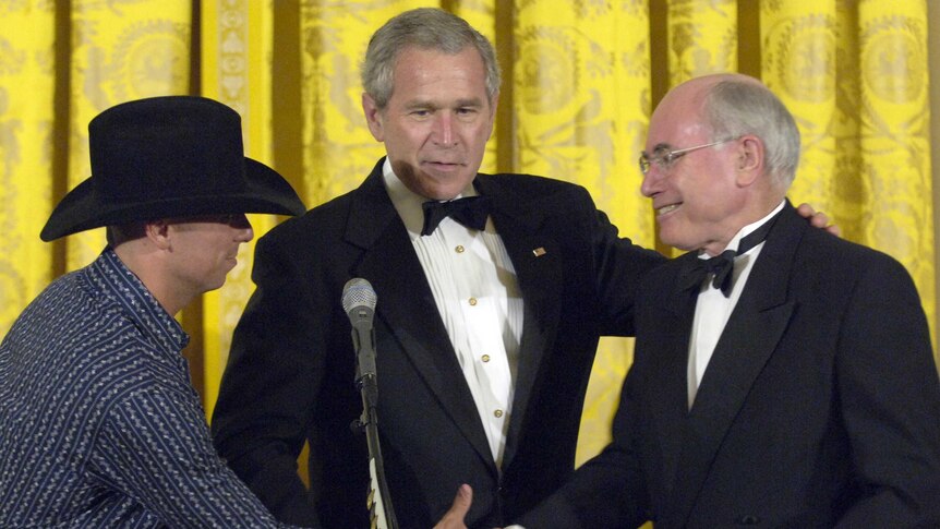 Two men in tuxedos shake hands with a man in a cowboy hat