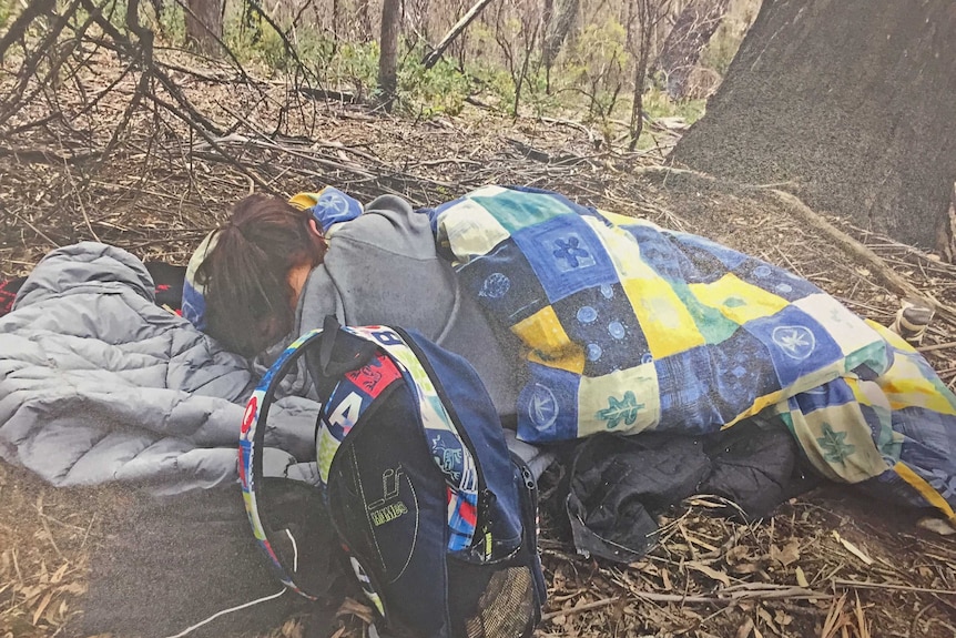 A woman appears to sleep under a blanket in bushland.