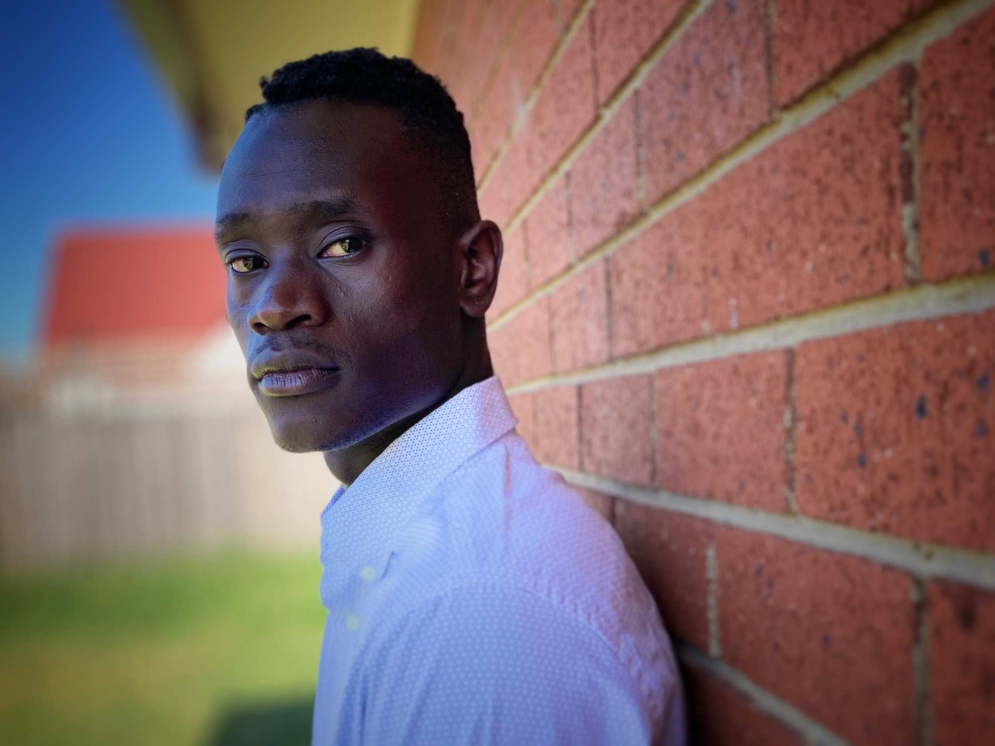 A young man leans against a brick wall looking into the camera.