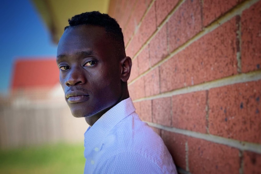 A young man leans against a brick wall looking into the camera.