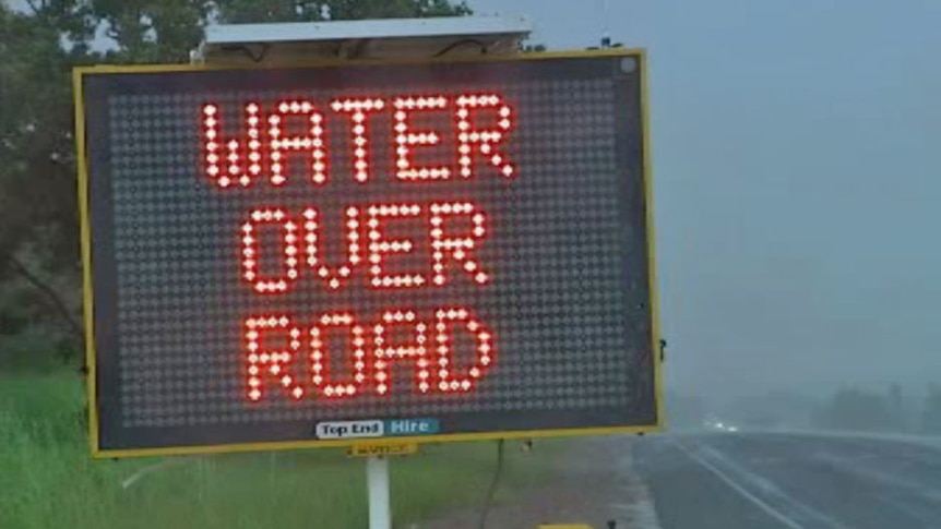A digital road sign displays the message WATER OVER ROAD. It appears to be heavily raining