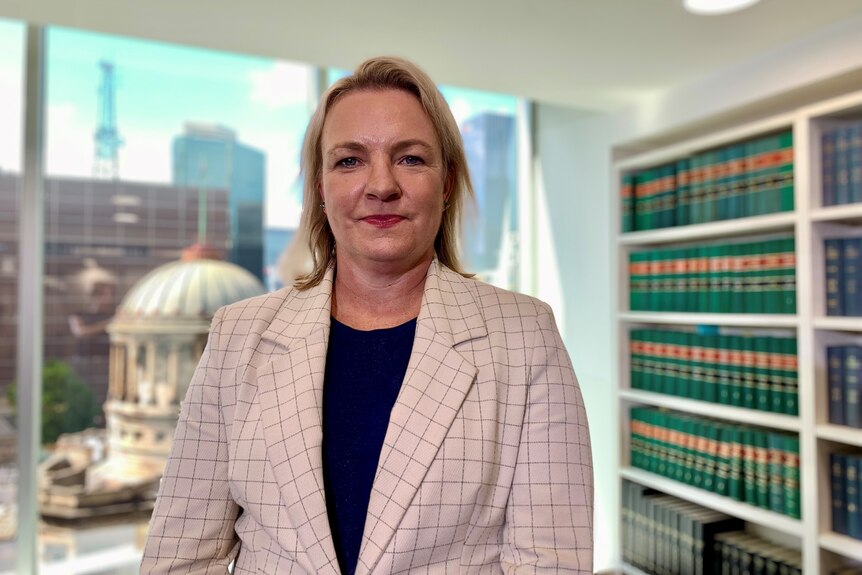 Sally Flynn wears a blazer and stands in front of a window with a view of Melbourne's supreme court