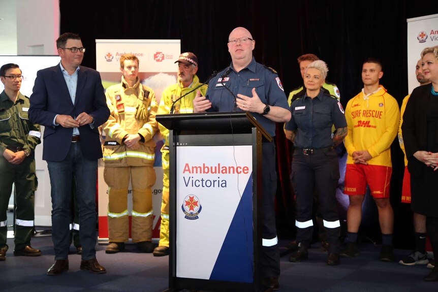 Tony Walker speaks at an 'Ambulance Victoria' podium as Daniel Andrews and first responders watch on.