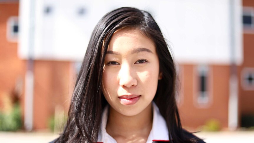Portrait of dark-haired, brown-eyed girl in school uniform, staring at camera in front of blurred school background.