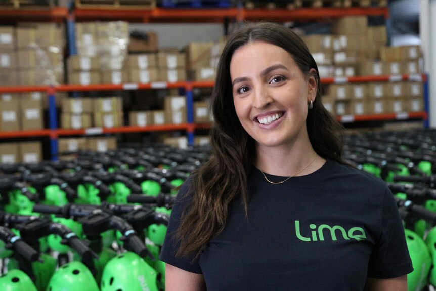 Woman with long brown hair wearing a black shirt with the Lime logo standing in front of e-scooters.
