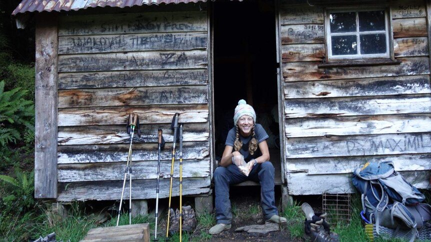 A smiling woman wearing beanie and casual clothes sits in the doorway of wooden hut with tin roof, next to a backpack.
