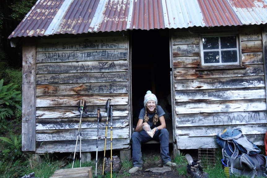 A smiling woman wearing beanie and casual clothes sits in the doorway of wooden hut with tin roof, next to a backpack.