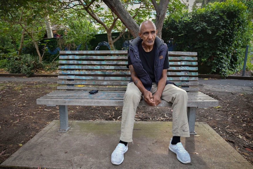 a man in a puffer vest sits dejectedly on a park benh