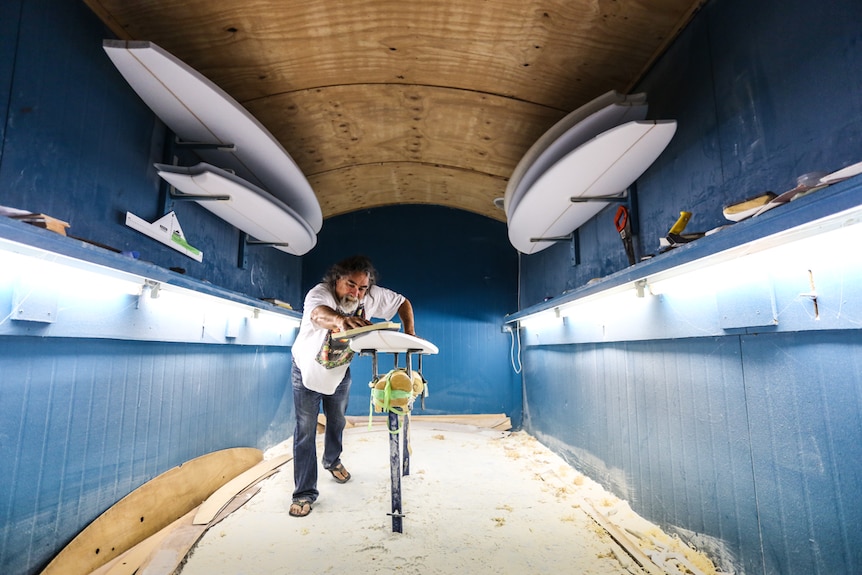 A man sands a surfboard inside a large shed workshop.