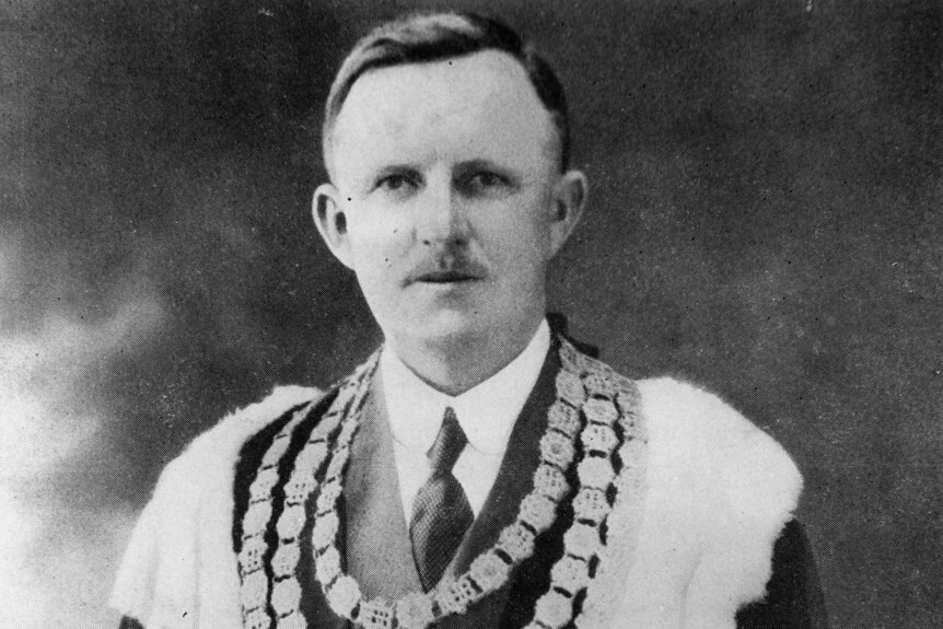 A black and white image of a man with a moustache wearing a tie and mayoral robes.