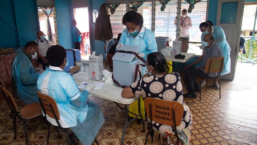 Three people sit at table in hospital scrubs and masks while woman stands in front.