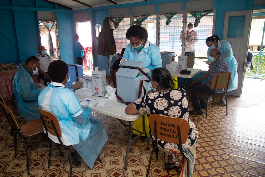 Three people sit at table in hospital scrubs and masks while woman stands in front.