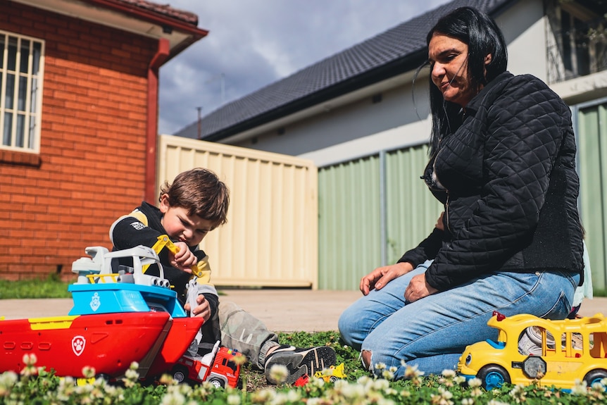 A boy plays with a toy while sitting on the grass in the backyard while his mother watches on.