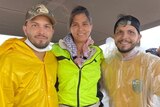 Dark haired woman stands in middle of two dark haired, bearded men, all wearing yellow jackets