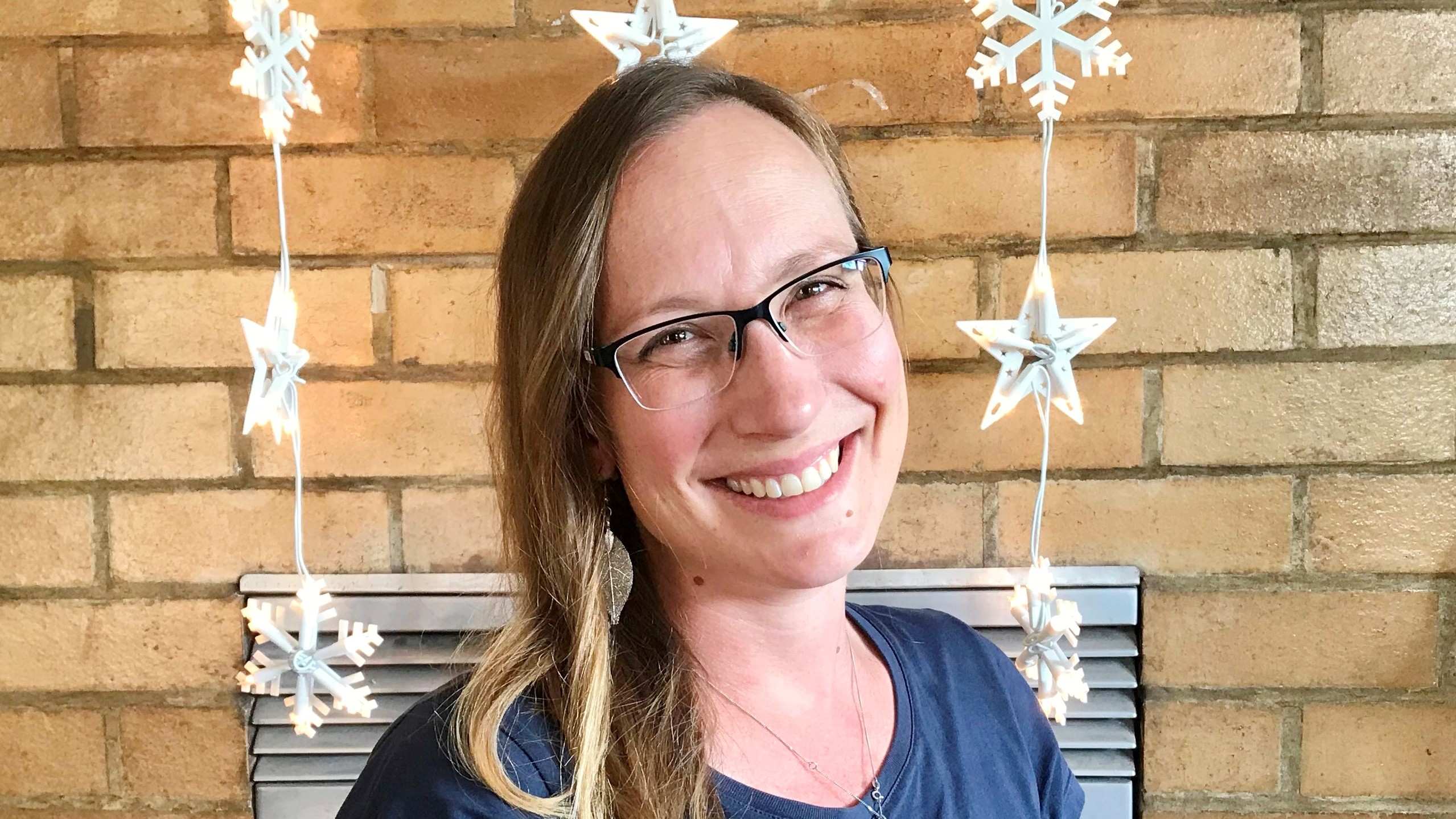 Photograph of a white woman with straight blond hair smiling at the camera. She is wearing glasses.