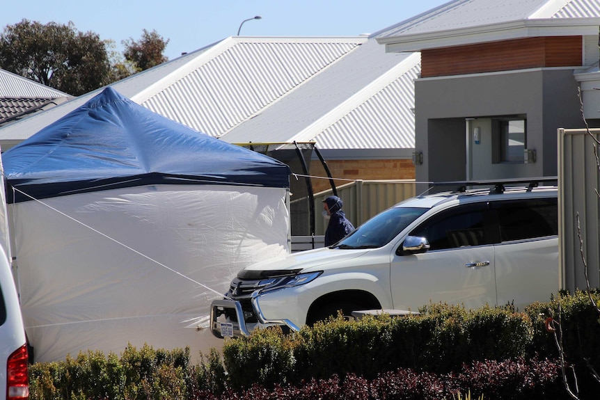 A tent and car outside a suburban home, with a police officer in a blue jumpsuit and mask in the front yard.