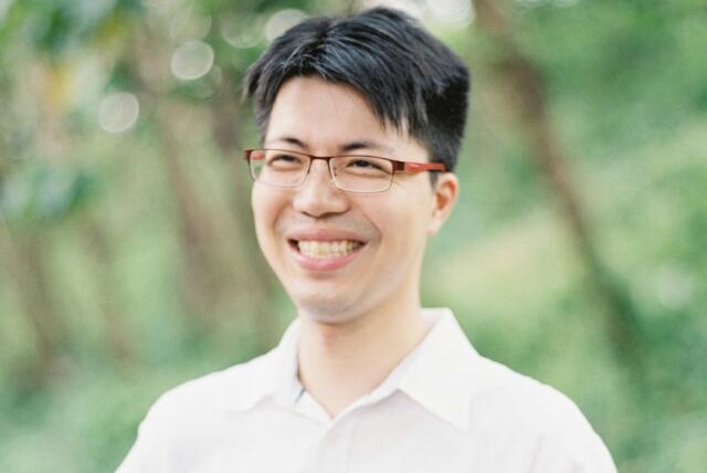 A smiling young man in a white shirt with trees in the background.