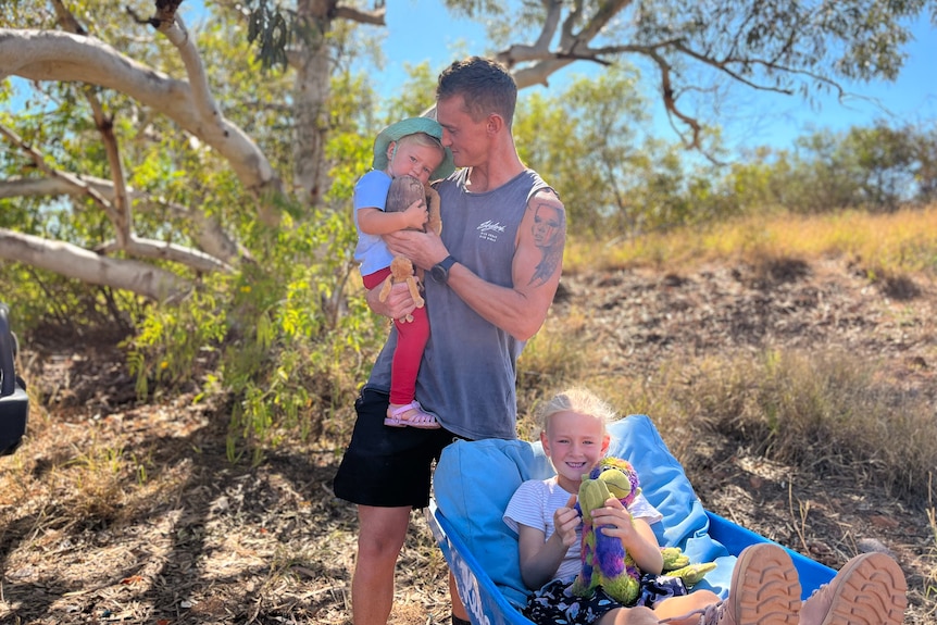 A man with tattoos holds his toddler daughter while his six-year-old daughter sits in a wheelbarrow.