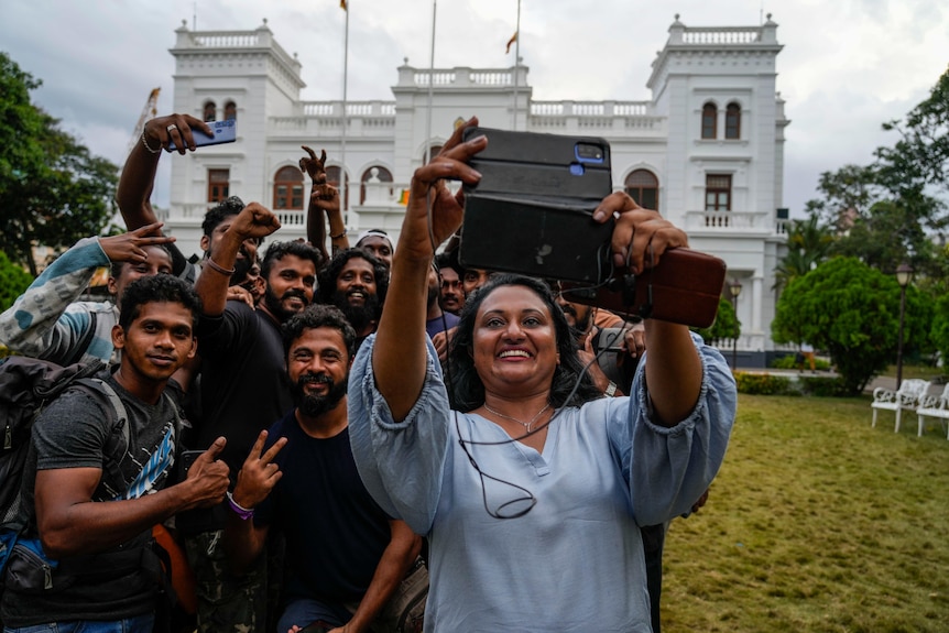 A woman holds up a phone as a group poses for a selfie in front of a white government building