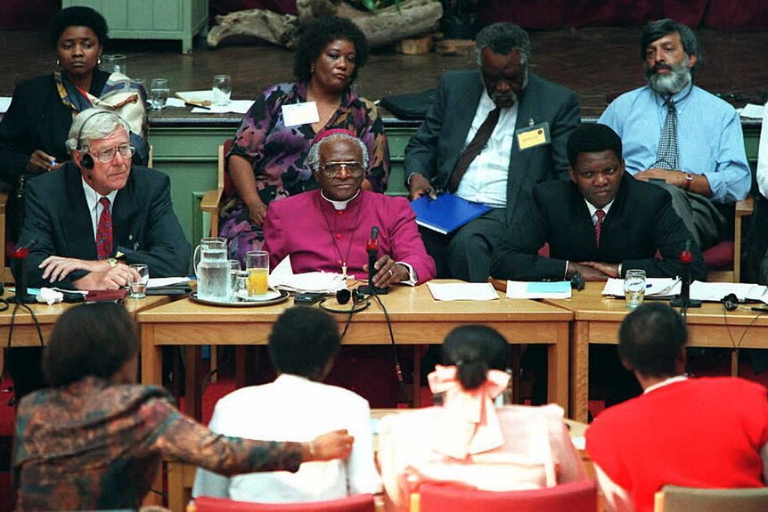 Desmond Tutu sits behind a desk in a purple habit surrounded by bureaucrats