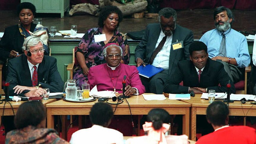 Desmond Tutu sits behind a desk in a purple habit surrounded by bureaucrats
