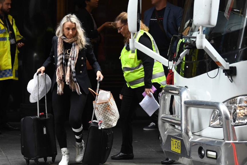 A woman with suitcases outside a hotel, with a man and a woman in hi-vis vests behind her. 