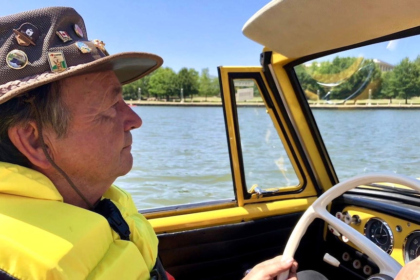 A man in a fishing hat sits behind the wheel of a car that is floating in a lake.