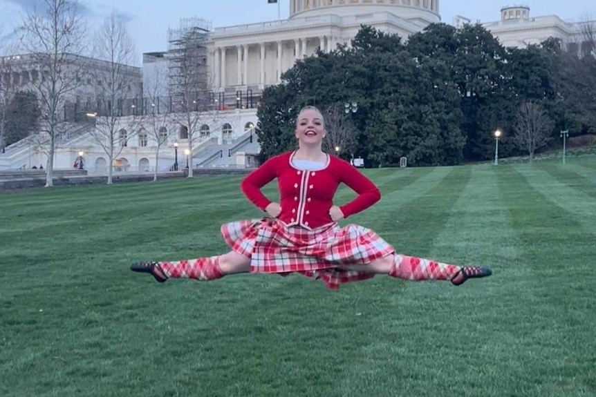 Tully Stone dressed in red top, white and red tartan skirt and socks, dancing in front of a white building.
