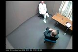 A still from police security camera footage shows two men in a police interrogation room.