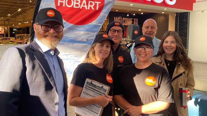 Six people are grouped together and smiling. They are wearing ABC Radio Hobart branded shirts and hats.
