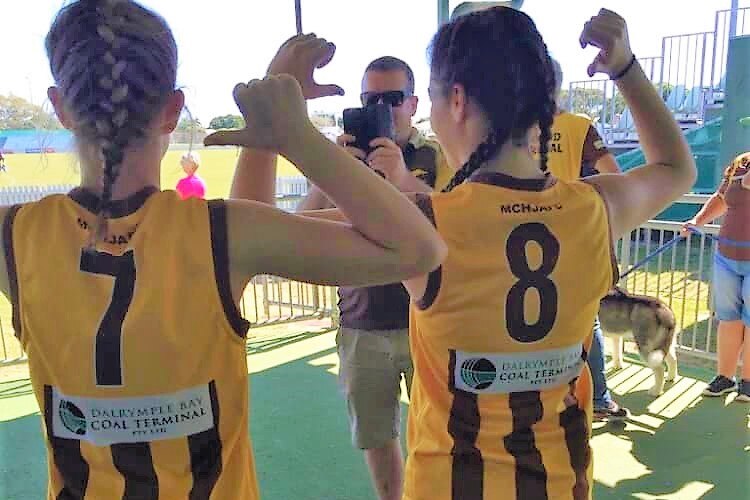 Two young female AFL players with their backs to the camera pointing at their jersey number.