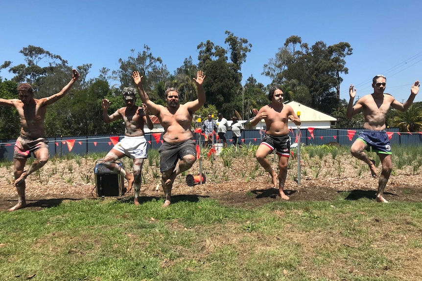 Five young men in traditional Indigenous body paint dance in front of a newly-planted garden.
