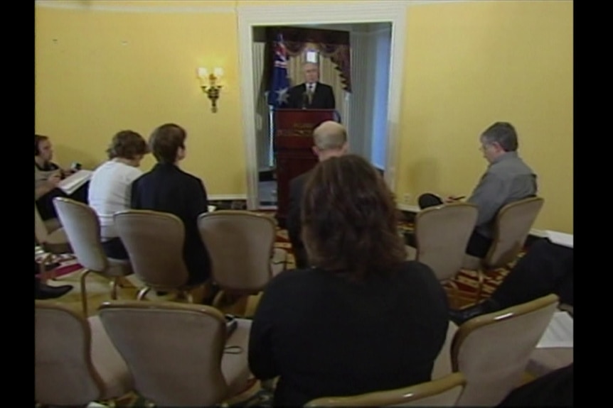 Howard behind lectern talking to journalists seated on chairs.
